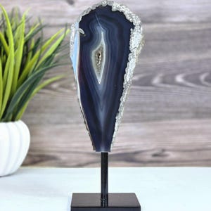 May include: A polished, dark blue agate geode slice mounted on a black metal stand. The geode features concentric bands of color, with a central crystalline cavity. The outer edge is lined with sparkling crystals, creating a decorative display piece.
