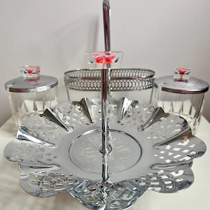 May include: A silver three-tiered serving set with a handle. The top tier is a round bowl with a decorative edge. The middle tier is a round plate with a decorative edge. The bottom tier is a round plate with a decorative edge. Each tier has a glass jar with a lid. The lids have a pink flower design.