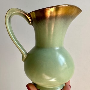 May include: A light green ceramic jug with a gold rim and handle. The jug has a rounded body and a flared lip.
