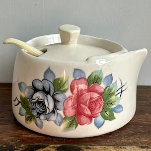 May include: A white ceramic sugar bowl with a lid and a matching spoon. The bowl is decorated with a floral pattern featuring pink and blue roses.
