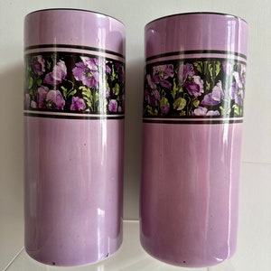 May include: Two tall, cylindrical ceramic vases with a lavender glaze. Each vase has a band of black and white trim with a floral pattern of purple poppies and green leaves.