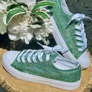 Op de afbeelding: Een paar lage sneakers bedekt met felgroene glitters met witte rubberen zolen, witte neuzen en witte veters. De schoenen hebben een textuurrijke, sprankelende afwerking over het bovenwerk en zijn gestrikt met platte witte veters.