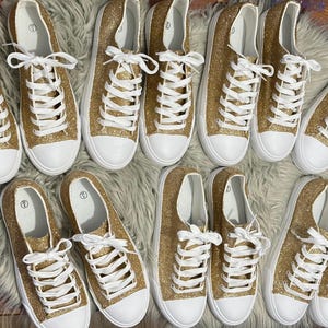 May include: A group of ten gold glitter trainers with white laces and white soles. The trainers are arranged in two rows of five.