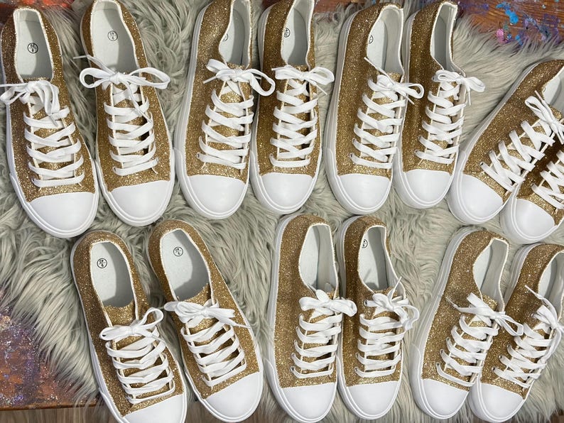 May include: A group of ten gold glitter trainers with white laces and white soles. The trainers are arranged in two rows of five.