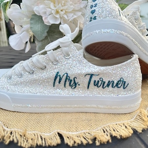 Puede incluir: Zapatillas blancas de brillantina con letras color turquesa que dicen "Mrs. Turner". El zapato derecho tiene un diseño de brillantina con corazones y números color turquesa.