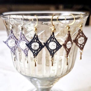 May include: Six handmade macrame earrings in a variety of colors, including purple, navy, brown, and black. Each earring features a diamond-shaped design with a clear, round bead in the center and a small gold-colored teardrop charm.