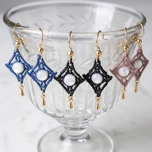 May include: A collection of handmade macrame earrings in various colors, including blue, black, and mauve. Each earring features a diamond-shaped design with a central white bead and gold-colored accents. The earrings are displayed on a glass bowl.