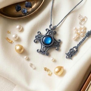 May include: A handcrafted, gray macrame necklace with a blue gemstone center, surrounded by a decorative design. The necklace is displayed with pearls, gold accents, and the text "N's Macrame".