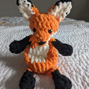 May include: A crocheted fox toy in orange, white, and black. The fox has large ears, a white chest, and black paws. The toy is placed on a white bedspread.