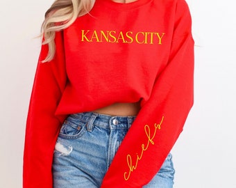 May include: Red cropped sweatshirt with the text "KANSAS CITY" in yellow on the front and "chiefs" in yellow script on the left sleeve.