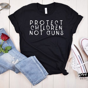 May include: Black t-shirt with white text that reads "PROTECT CHILDREN NOT GUNS". The shirt is laid out on a white wooden surface with a pair of blue jeans, a red rose, and white sneakers.