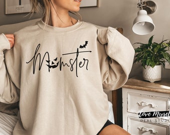 May include: A beige sweatshirt featuring the word "Monster" in black script. The letter "o" in "Monster" is replaced with a jack-o'-lantern face, and a bat graphic is above the "r". This is a Halloween-themed garment.