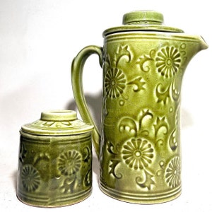 May include: A vintage green ceramic coffee pot and sugar bowl set. The set features a floral pattern and a crackle glaze finish. The coffee pot has a handle and a lid. The sugar bowl has a lid. The set is a light green colour.