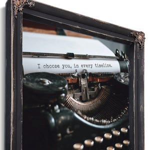 May include: Framed print featuring a vintage typewriter with the text "I choose you, in every timeline." The typewriter is black with silver keys and a paper sheet. The frame is black with ornate corner details.