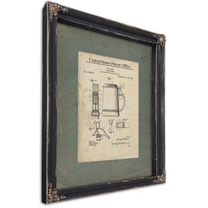 May include: Framed patent print featuring a detailed illustration of a beer stein design. The print is from the United States Patent Office and is encased in a black wooden frame with ornate corner accents. The background is a muted green.
