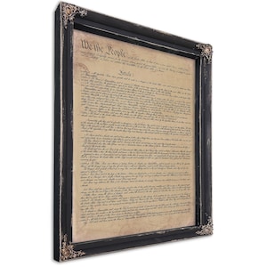 May include: Framed print of the U.S. Constitution. The document is displayed in a black wooden frame with ornate corner details. The text "We the People" is visible at the top. The paper has a vintage, aged appearance.