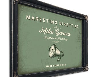 May include: A rectangular wall sign in a distressed black frame with ornate corner details. The sage green center features the text MARKETING DIRECTOR, Mike Garcia, Brightside Marketing, and MAKE SOME NOISE, with a megaphone illustration.