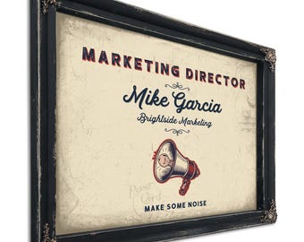 May include: A rectangular framed sign with a distressed black finish and ornate corner details. The cream-colored center features the text MARKETING DIRECTOR, Mike Garcia, Brightside Marketing, and MAKE SOME NOISE, with a megaphone illustration.