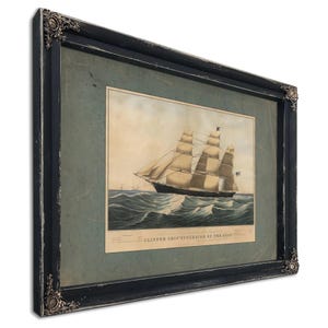 May include: Framed print of a clipper ship sailing on the ocean. The artwork features a detailed illustration of the ship with billowing sails, set against a backdrop of waves and a cloudy sky. The frame is black with ornate corner details.