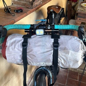 May include: A bicycle handlebar with teal tape, a light, and a light gray bag secured with black straps. The bag has the word "ORCEATTI" printed on it. The bike is in front of a window and a table.