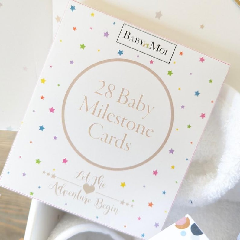 May include: A white box of 28 Baby Milestone Cards with a star pattern. The box has a circular design with the text "28 Baby Milestone Cards" and "Let The Adventure Begin". The brand name "Baby Moi" is at the top.