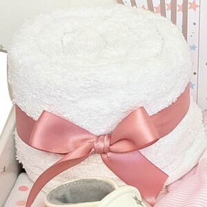 May include: A rolled white towel tied with a pink satin ribbon and bow. The towel is part of a baby gift set, with a glimpse of a baby shoe and other items in the background. The set is presented in a gift box.