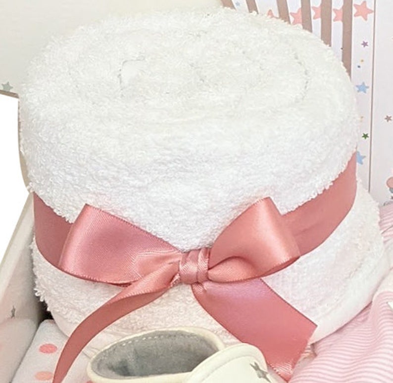 May include: A rolled white towel tied with a pink satin ribbon and bow. The towel is part of a baby gift set, with a glimpse of a baby shoe and other items in the background. The set is presented in a gift box.