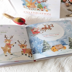 May include: A children's book titled "Celebration" with illustrations of reindeer, Santa Claus, and a fox in a snowy scene. The open pages display colorful images and text, with a decorative bird figurine in the foreground.