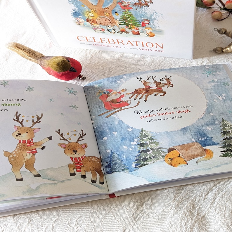 May include: A children's book titled "Celebration" with illustrations of reindeer, Santa Claus, and a fox in a snowy scene. The open pages display colorful images and text, with a decorative bird figurine in the foreground.