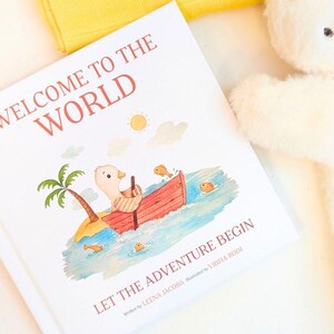 May include: A children's book titled "Welcome to the World" with an illustration of a duck in a boat. The book cover features a palm tree, sun, and the text "Let the Adventure Begin". A plush duck toy is in the background.