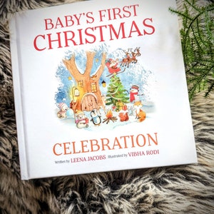 May include: A hardcover children's book titled "Baby's First Christmas Celebration" with illustrations of a winter scene. The cover features a tree with a door, a Christmas tree, Santa Claus, and various animals. The author is Leena Jacobs, and the illustrator is Vibha Rodi.
