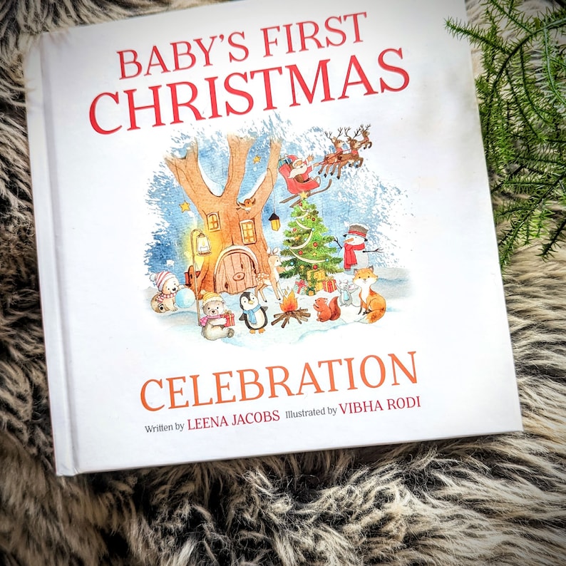 May include: A hardcover children's book titled "Baby's First Christmas Celebration" with illustrations of a winter scene. The cover features a tree with a door, a Christmas tree, Santa Claus, and various animals. The author is Leena Jacobs, and the illustrator is Vibha Rodi.