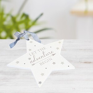 May include: A white star-shaped ornament with gray star accents and the text "LET THE adventure BEGIN" in gray. A gray ribbon is attached at the top. The ornament is on a light wood surface.