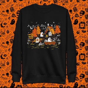 Puede incluir: Sudadera negra con un gráfico de fantasmas acampando junto a un lago por la noche. El gráfico incluye una fogata, una tienda de campaña y un cartel que dice "Camp Spooky Lake".