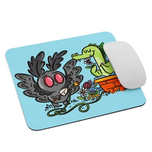 May include: A blue mousepad with a cartoon illustration of a black moth with red eyes holding a watering can and a green alligator in a flower pot. The moth is watering flowers.