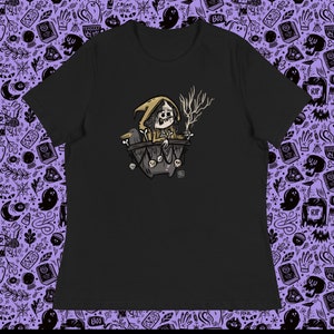 Könnte beinhalten: Schwarzes T-Shirt mit einem Grafikdruck einer Hexe in einem Kessel mit einer Krähe und einer skelettartigen Hand. Die Hexe trägt eine braune Kapuzenrobe und hat eine lange, spitze Nase.