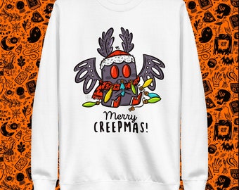 Könnte beinhalten: Weißer Pullover mit einer Cartoon-Illustration einer Fledermaus, die einen Weihnachtsmann-Hut trägt und Weihnachtslichter hält. Der Text "Merry Creepmas!" ist unter der Fledermaus gedruckt.