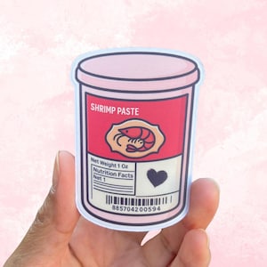 May include: A sticker featuring a pink and red container of shrimp paste. The label reads "SHRIMP PASTE" and includes a shrimp illustration, a heart, and a barcode. Net weight is 1 oz.