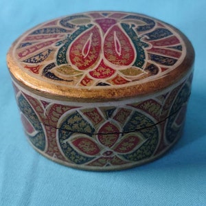 May include: A round, decorative box with a gold, red, green, and blue floral pattern. The box is made of paper mache and has a hinged lid.