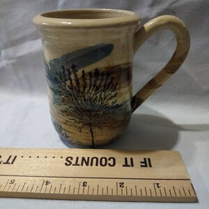 May include: A beige ceramic mug with a brown tree and blue sky design. The mug has a handle and the words "If It Counts" are printed on the bottom of the mug.