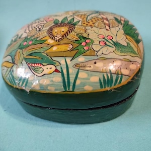 May include: A green oval-shaped trinket box with a hinged lid. The lid is painted with a whimsical scene of animals and plants in a jungle setting. The animals include a lion, a bird, and a fish. The colors are bright and vibrant, including green, yellow, blue, and pink.