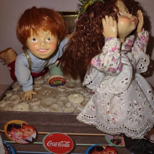May include: A vintage Coca-Cola tin box with a brown and white striped design. The box is filled with buttons and features two dolls, a girl with a floral crown and a boy with red hair. The girl is wearing a white dress with lace trim and the boy is wearing a blue shirt and red pants.