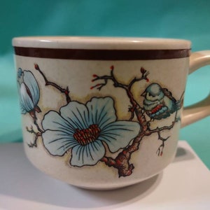 May include: A beige ceramic mug with a brown rim and a floral design featuring blue flowers and birds. The mug has a handle.