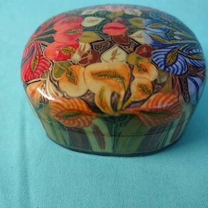May include: A round, colorful trinket box with a floral design. The box is made of papier-mâché and features a variety of flowers in shades of red, orange, yellow, blue, and green.