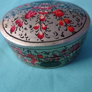 May include: A round, silver-colored box with a floral design. The lid is painted with red and white flowers on a silver background. The box is about 3 inches in diameter.