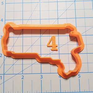 Puede incluir: Un cortador de galletas de plástico naranja con forma de bocadillo con el número 4. El cortador de galletas mide aproximadamente 12,7 cm de largo y 7,6 cm de ancho.