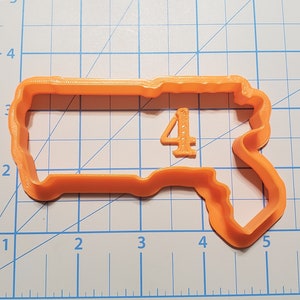 Puede incluir: Un cortador de galletas de plástico naranja con forma del número 4 y una forma rectangular con borde ondulado. El número 4 es pequeño y tiene una fuente decorativa. El cortador está sobre una cuadrícula azul y blanca.