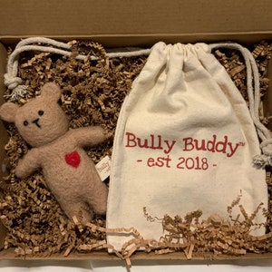 Könnte beinhalten: Ein brauner Filzteddybär mit einem roten Herzen auf der Brust befindet sich in einer Geschenkbox mit braunem Papierstreu. Die Box enthält auch eine weiße Stofftasche mit dem Text "Bully Buddy - est 2018 -" in rot.