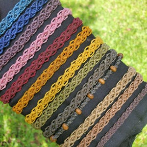 Peut inclure: Une collection de dix bracelets en macramé disposés en rangée diagonale. Les bracelets présentent diverses couleurs dont bleu, violet, rose, rouge, jaune, vert olive et marron. Un bracelet gris est orné de petites perles en bois marron clair.
