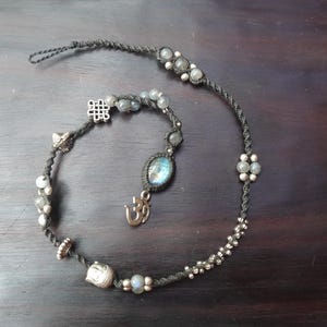 Peut inclure: Un collier artisanal avec un cordon foncé, orné de perles, de breloques et de pierres précieuses argentées. Le collier comporte un pendentif central avec une pierre bleue et un symbole Om, ainsi que d'autres éléments décoratifs. Le design est complexe et bohème.